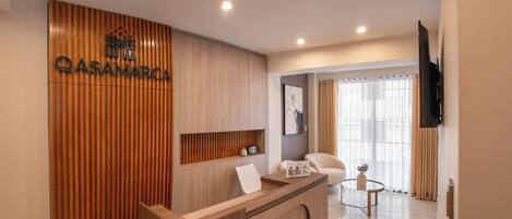 Un área de recepción moderna con un mostrador de madera, una televisión montada y una zona de estar con una mesa de vidrio.