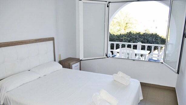 A neatly made bed with white linens and two rolled towels, a small bedside table, and a window with a view of a white balcony and greenery outside. - Anacasa San Fernando MET Oliva Nova AP611 (Oliva)