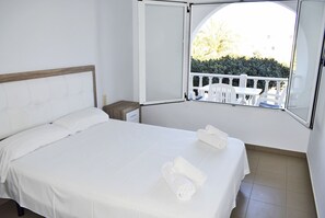 Una cama bien tendida con sábanas blancas y dos toallas enrolladas, una mesita de noche pequeña, y una ventana que da a un balcón blanco y follaje exterior.