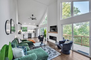 Ein geräumiges Wohnzimmer mit einer grünen Sofagarnitur, einem Couchtisch und einem Kamin. Im Hintergrund ist eine Küche mit einem großen Fenster mit Blick auf eine Terrasse zu sehen.