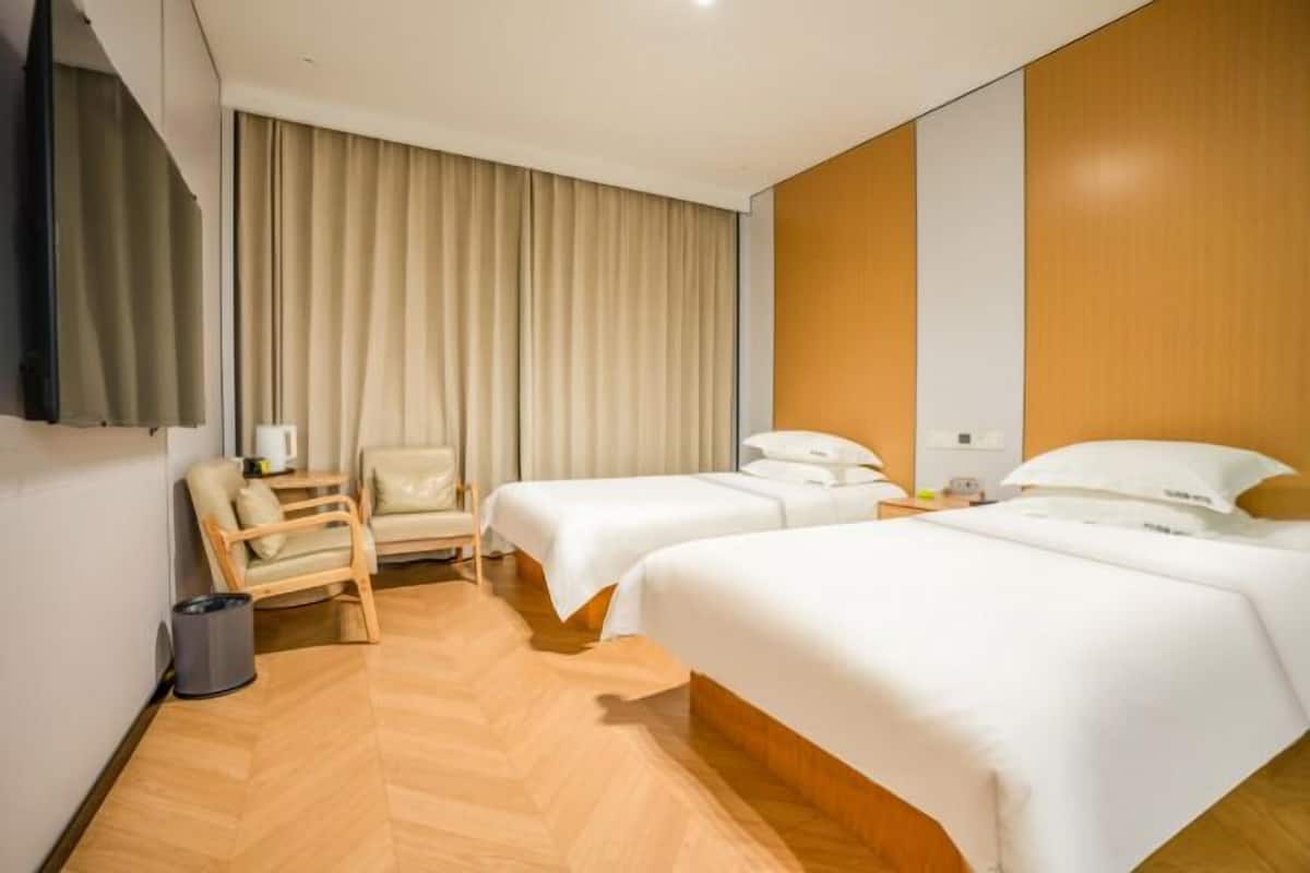 Quarto de hotel com duas camas, uma cadeira, uma televisão e piso de madeira.