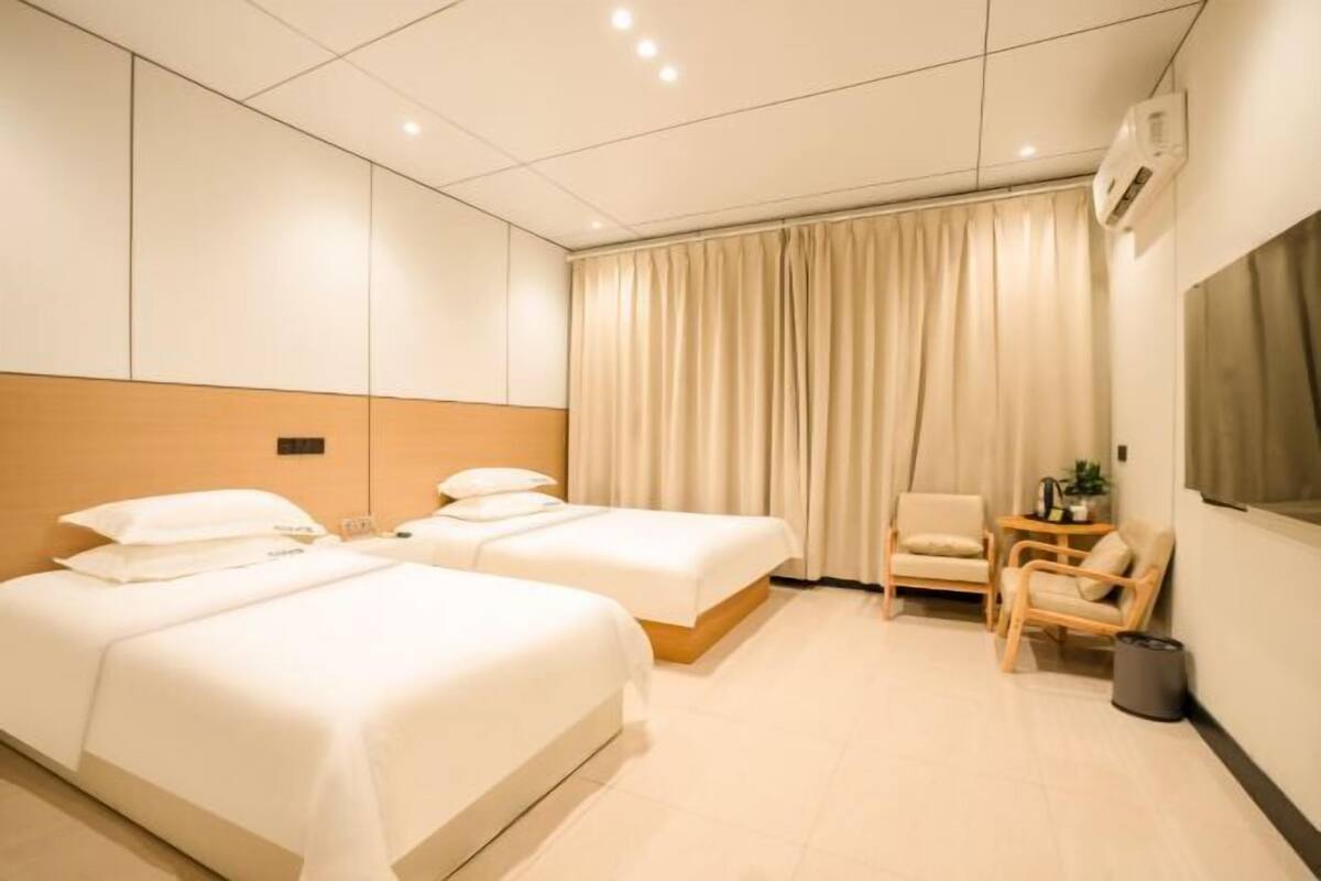 Quarto de hotel com duas camas, uma cadeira, uma mesinha e uma janela com cortinas.