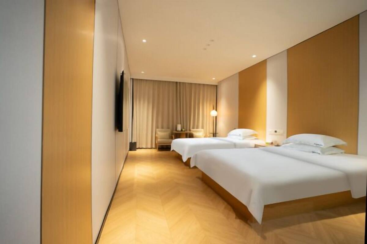 Quarto de hotel com duas camas, uma televisão de tela plana fixada na parede, uma mesinha de cabeceira com abajur e uma janela grande com cortinas.