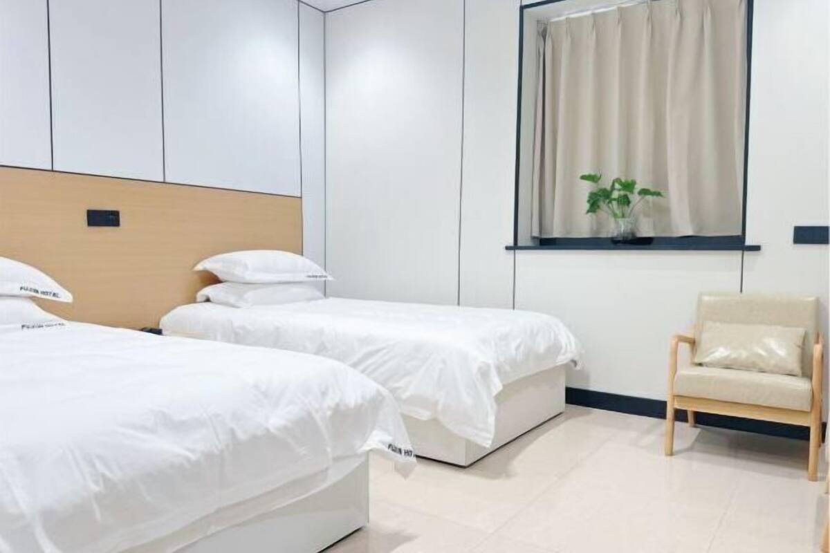 Quarto de hotel com duas camas de solteiro, cabeceira de madeira, janela com cortinas, uma planta pequena e uma cadeira de madeira.