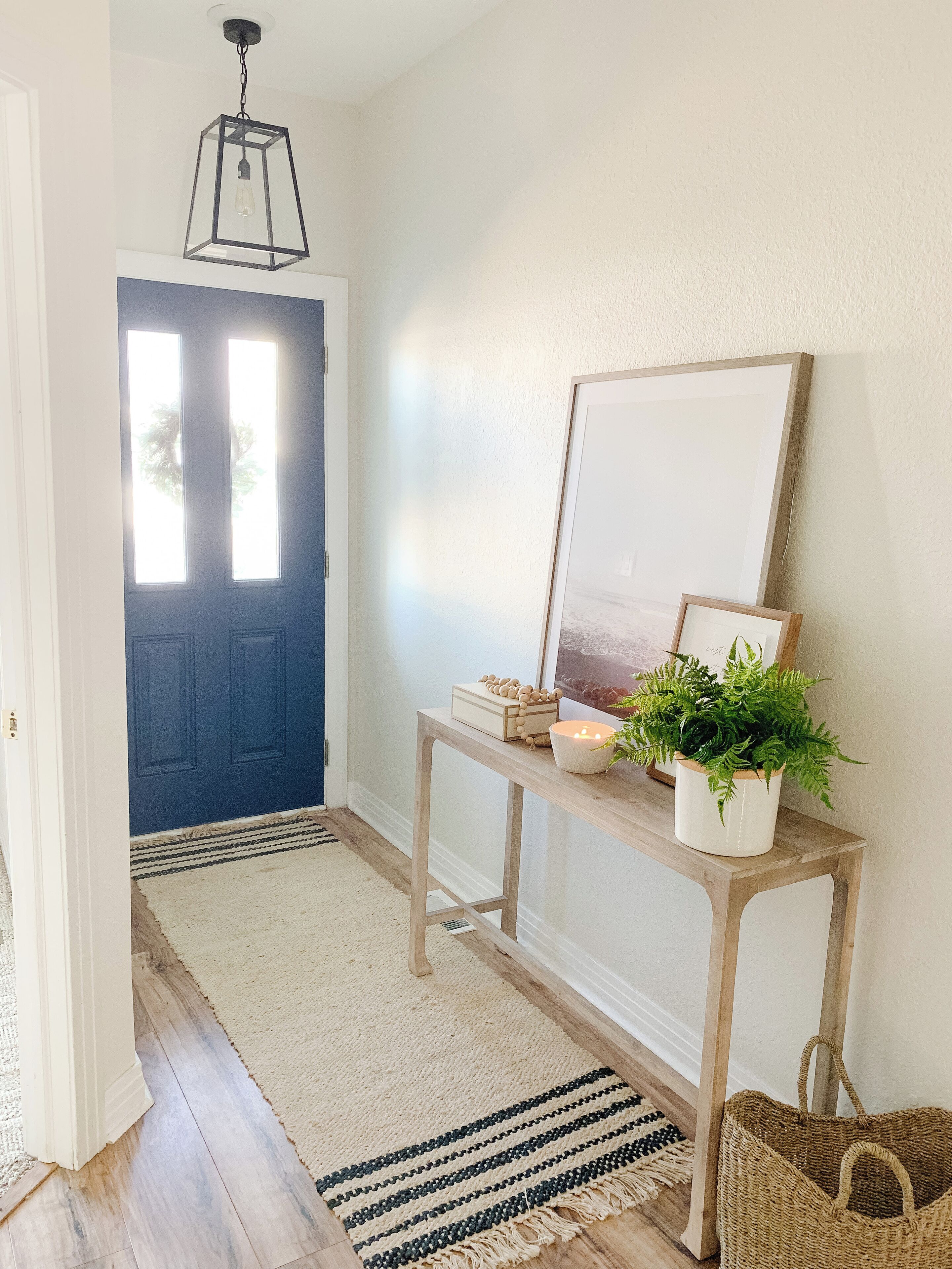Un pasillo con una puerta azul, una consola de madera con una planta, velas y un cuadro enmarcado.