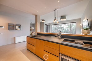 A modern kitchen with a large island, pendant lights, and a view of trees through a large window.