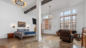 Un'ampia stanza con un letto grande, un divano in pelle e un tavolo da pranzo. Sono presenti diverse finestre con tende, un lampadario e una televisione a parete.