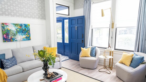 A modern living room with a grey sofa, a round coffee table, and two armchairs with yellow and blue cushions. A blue door and a large window with curtains are visible.