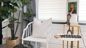 A room with a white chair, a large green plant, a white cushion, a decorative bowl, and a small table with a black and white object.