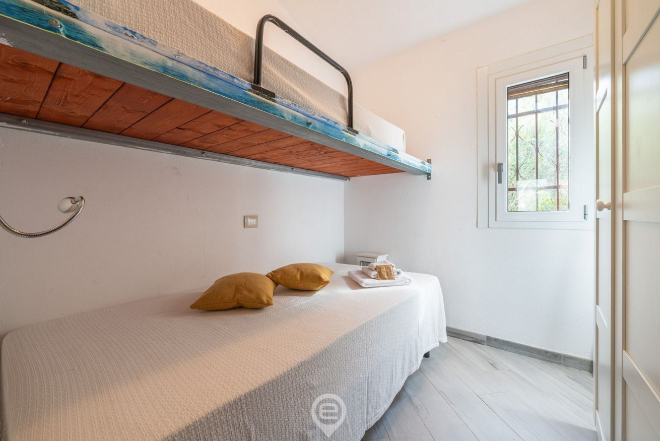 Habitación individual con una cama nido superior, ventana con vistas al verdor y una pequeña mesa con una bandeja de pan y mantequilla.