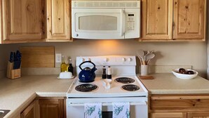 Una cocina con armarios de madera, un microondas blanco, una estufa con un hervidor y diversos utensilios de cocina.