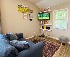 Ein Wohnzimmer mit einem blauen Sofa, einem Glastisch, einem an der Wand befestigten Fernseher und einem Fenster mit Jalousien.