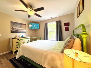 A bedroom with a bed, a ceiling fan, a television, a window with curtains, a dresser, and a lamp.