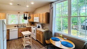 Una cocina con armarios de madera, una mesa de comedor con cojines azules y vistas a un jardín a través de grandes ventanales.