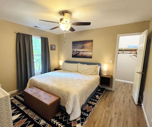 A bedroom with a bed, a ceiling fan, a nightstand, a bench, a painting, and a door. - Charming apartment in Pigeon Forge!!! Walk to the Alpine Coaster in minutes! (Pigeon Forge)