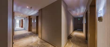 A hotel corridor with a patterned carpet, doors on the right, and a sign indicating the elevator location.