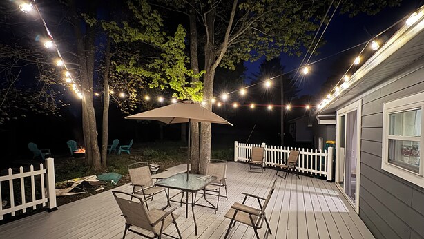Una terraza al aire libre con una mesa, sillas, luces colgantes y una valla de piquetes blancos.