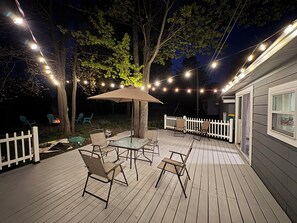 An outdoor deck with a table and chairs, string lights, and a white picket fence. - “Spacious 3BR Retreat with Deck, hot tub & Grill – Near Downtown Traverse City” (Traverse City)