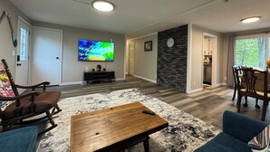 Una sala de estar moderna con un televisor de pantalla plana, una mesa de centro de madera, un tapete estampado y una zona de comedor con una pared de ladrillo decorativa y un reloj.