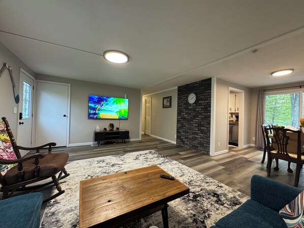 A modern living room with a flat-screen TV, a wooden coffee table, a patterned rug, and a dining area with a brick accent wall and a clock. - “Spacious 3BR Retreat with Deck, hot tub & Grill – Near Downtown Traverse City” (Traverse City)