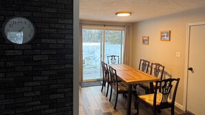 Un comedor con una mesa y sillas de madera, un reloj en la pared y vistas a un paisaje nevado a través de la puerta.