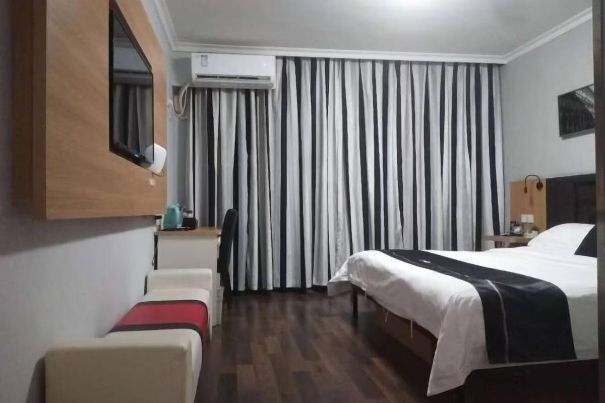 A hotel room with a bed, a desk, a chair, a TV mounted on the wall, and a window with curtains.