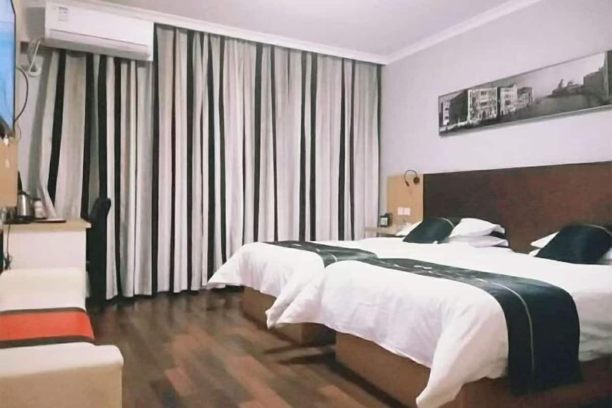 A hotel room with two beds, a desk, and a chair. There is a window with curtains, a wall-mounted air conditioner, and a framed picture on the wall.
