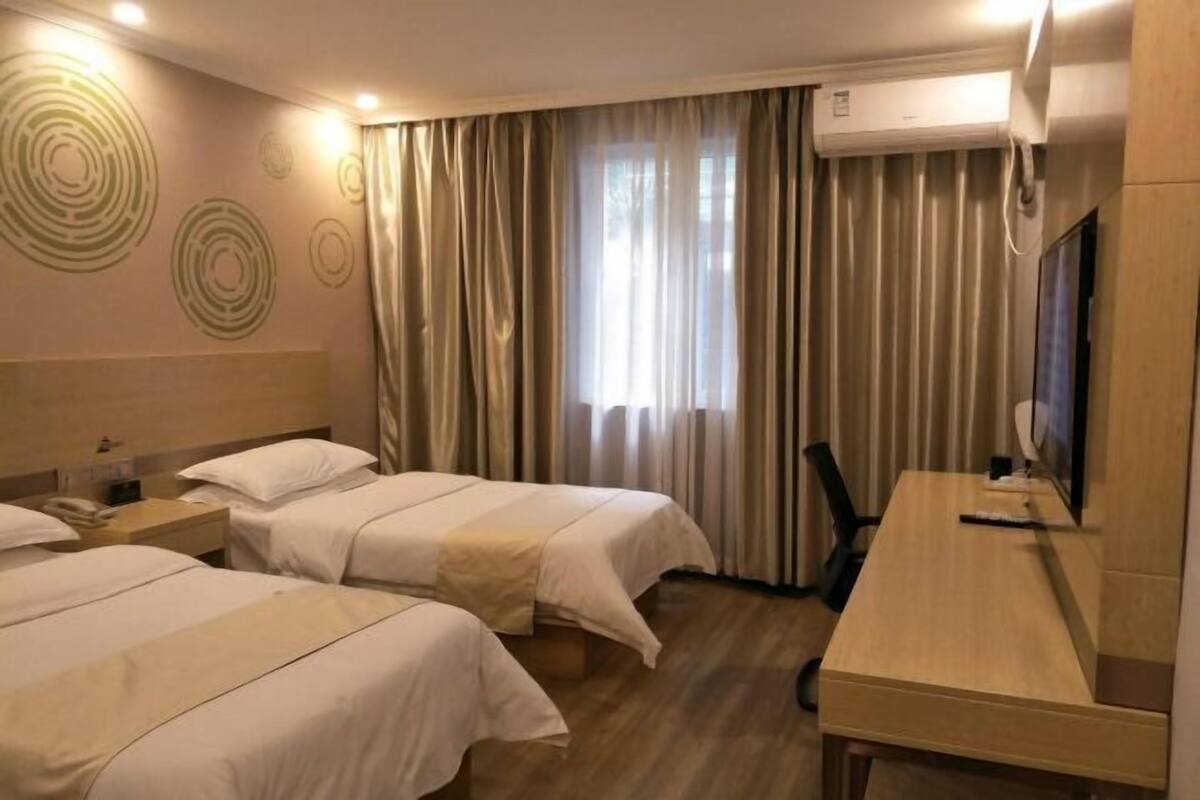 A hotel room with two beds, a desk, a TV, and an air conditioning unit.