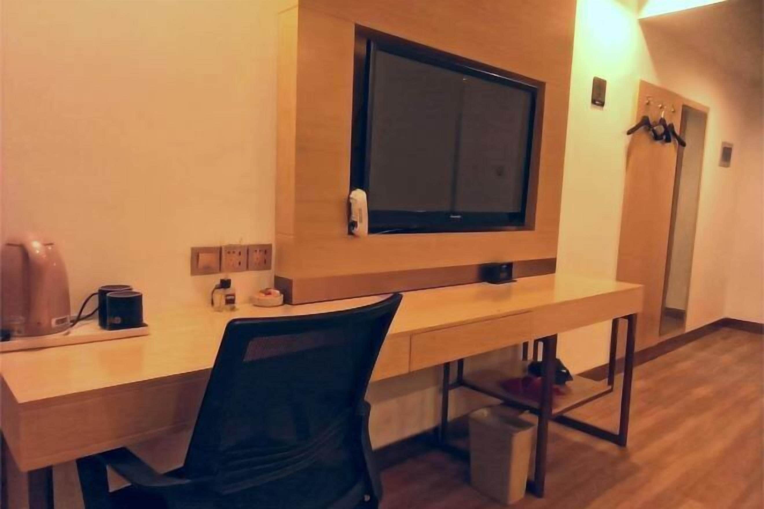 A hotel room with a wooden desk, a black office chair, a mounted television, and a coat rack.
