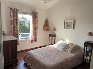 A bedroom with a bed, a dresser, a window with curtains, a bedside table with a lamp, and a framed picture on the wall. - 15 mn Bordeaux center near Bassin Arcachon St-Émilion ocean swimming SPA garden (Bruges)