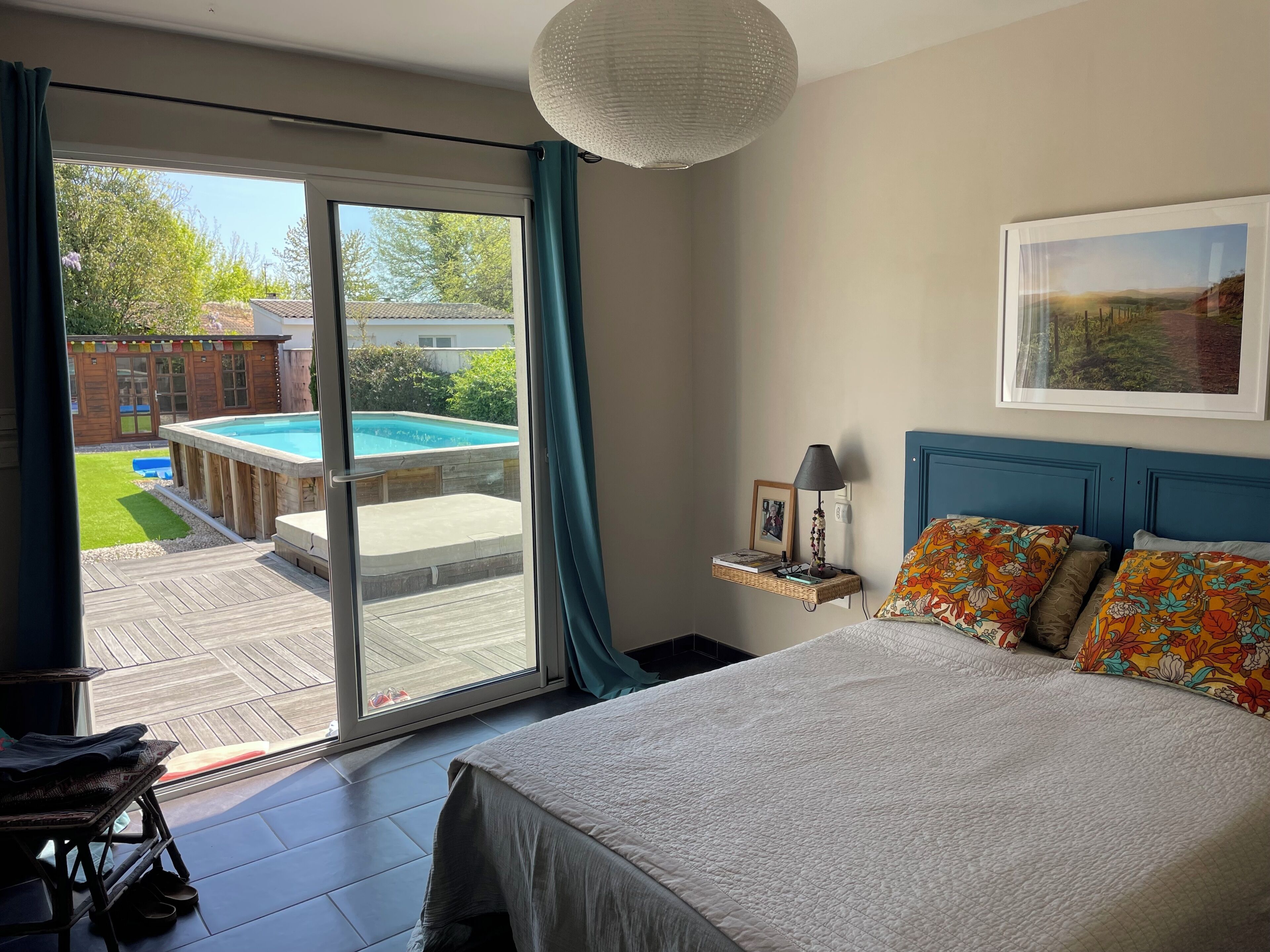 A bedroom with a bed, a nightstand, a lamp, a framed picture, a window with curtains, and a view of a swimming pool and garden.