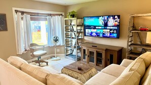 A living room with a sofa, a small table, a fan, a TV displaying a sports event, and a bookshelf. - Central Encinitas, 1.4 Miles from Beach (Encinitas)