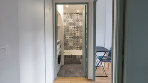 A narrow hallway leading to a bathroom with patterned tiles, a washing machine, and a small seating area with chairs.