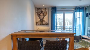 A dining area with a wooden table, two black chairs, and a green chair. There is a painting on the wall and a window with blue curtains.