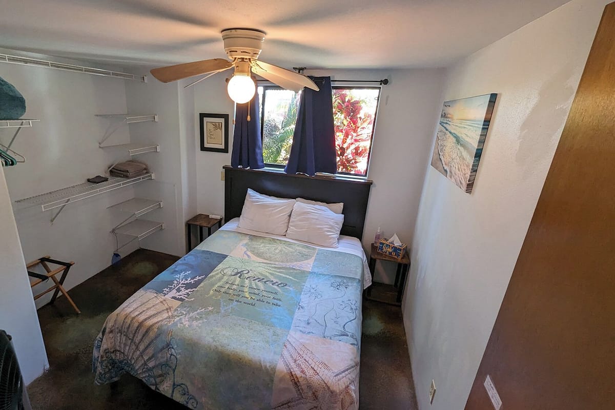 Ein Schlafzimmer mit einem Bett, einem Deckenventilator, einem Fenster mit Vorhängen, einem Regal und einem gerahmten Bild an der Wand.