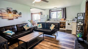 A modern living room with a sofa set, a coffee table, and a dining area with a table and chairs. There is a kitchen area with a stove and refrigerator.