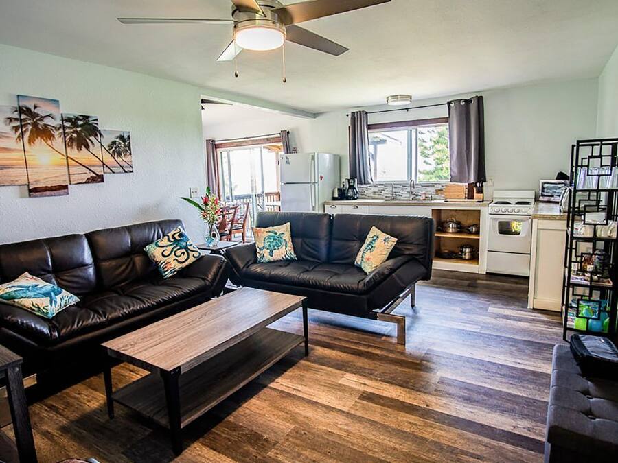 Una sala moderna con un conjunto de sofás, una mesa de centro y un comedor con mesa y sillas. Se observa también una zona de cocina con estufa y refrigerador.