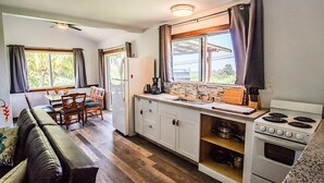 A compact kitchen with a dining area, a refrigerator, a stove, and a window with curtains. - Gods Peace Of Maui (Makawao)