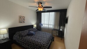 A hotel room with a bed, a ceiling fan, a nightstand with a lamp, a small table with a lamp, and a window with curtains.