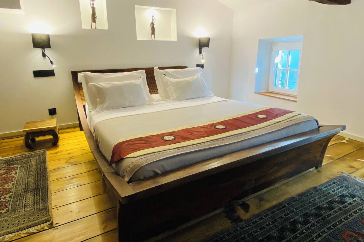 A bedroom with a wooden bed, white bedding, a red and beige blanket, two wall-mounted lights, a small wooden nightstand, a rug, and a window with a view.