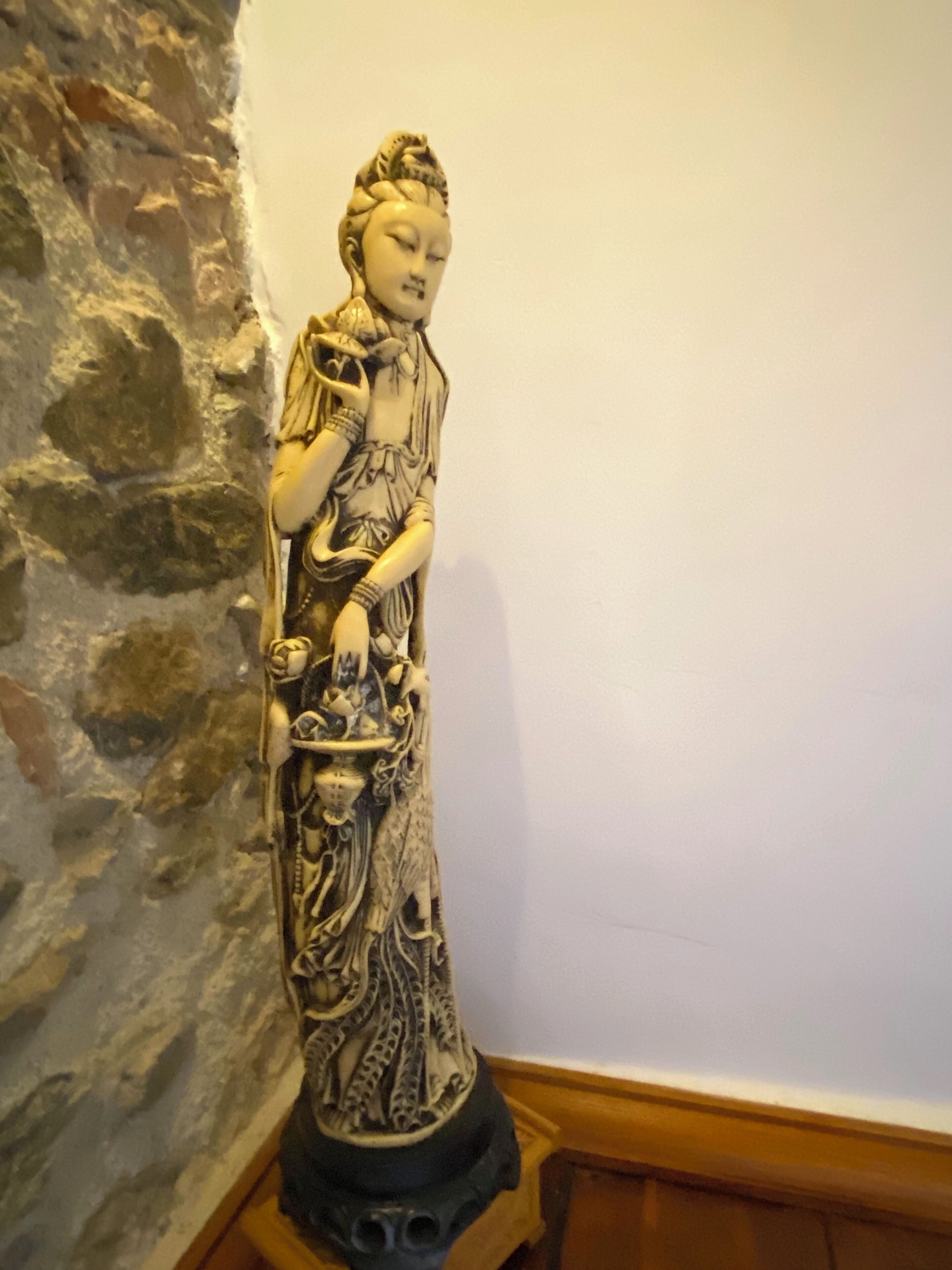Una estatua de una figura vestida con detalles intrincados, sobre un pedestal frente a un muro de piedra.