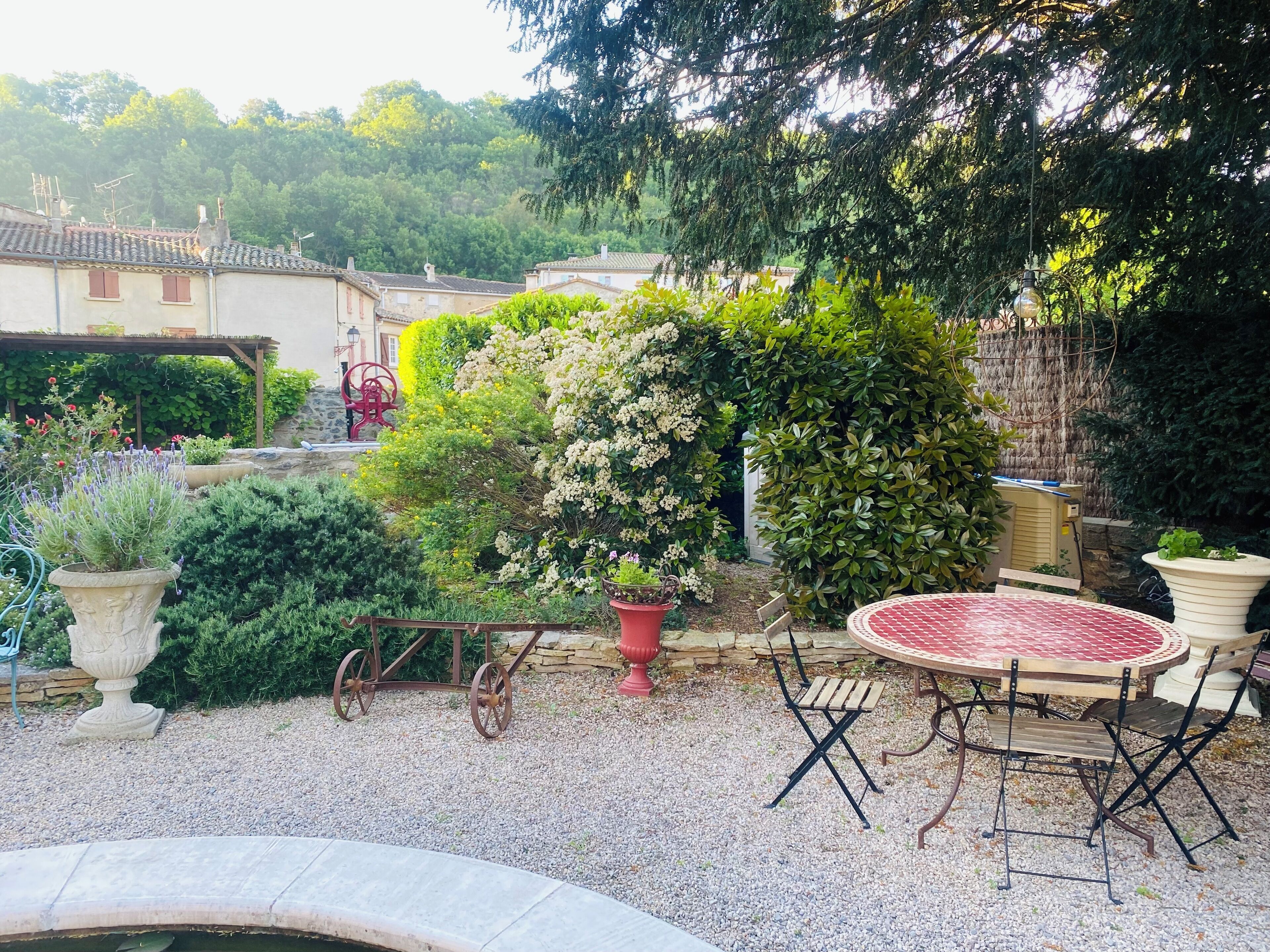 Une terrasse de jardin avec une table ronde et des chaises, entourée de plantes en pot et bordée d’un chemin de gravier.