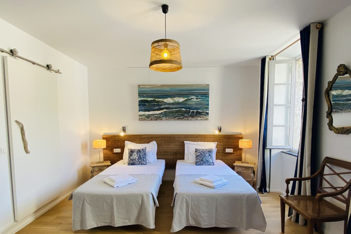 A hotel room with two beds, a painting of the sea, a wooden chair, and a window with curtains.