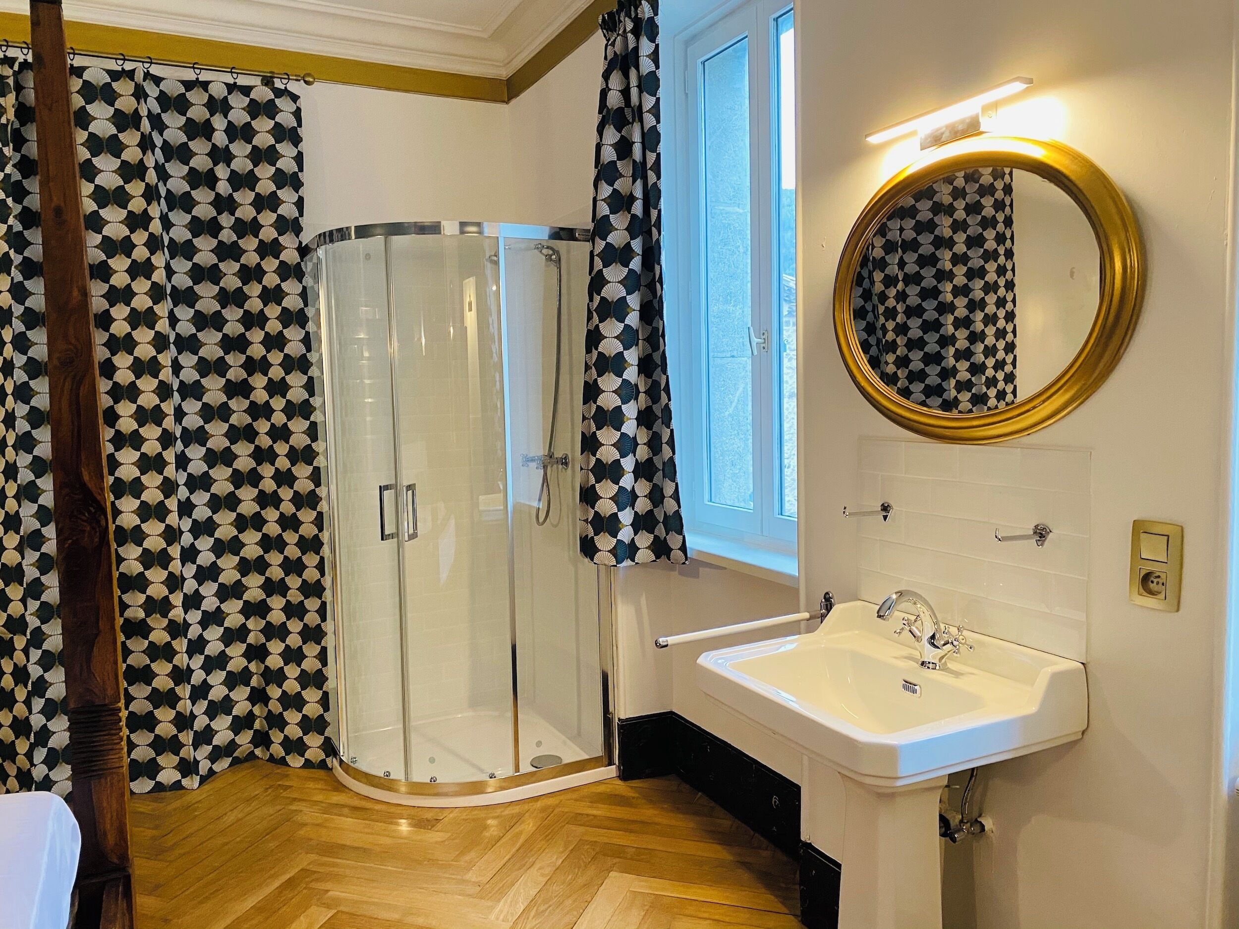 A bathroom with a glass shower enclosure, a white sink, a mirror with a gold frame, and patterned curtains.