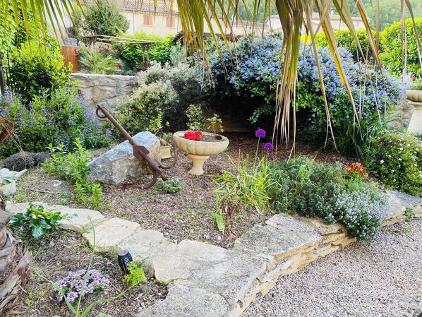 A garden with a rustic stone planter, a metal watering can, and various plants. - Aux Berges du Libre (Félines-Termenès)