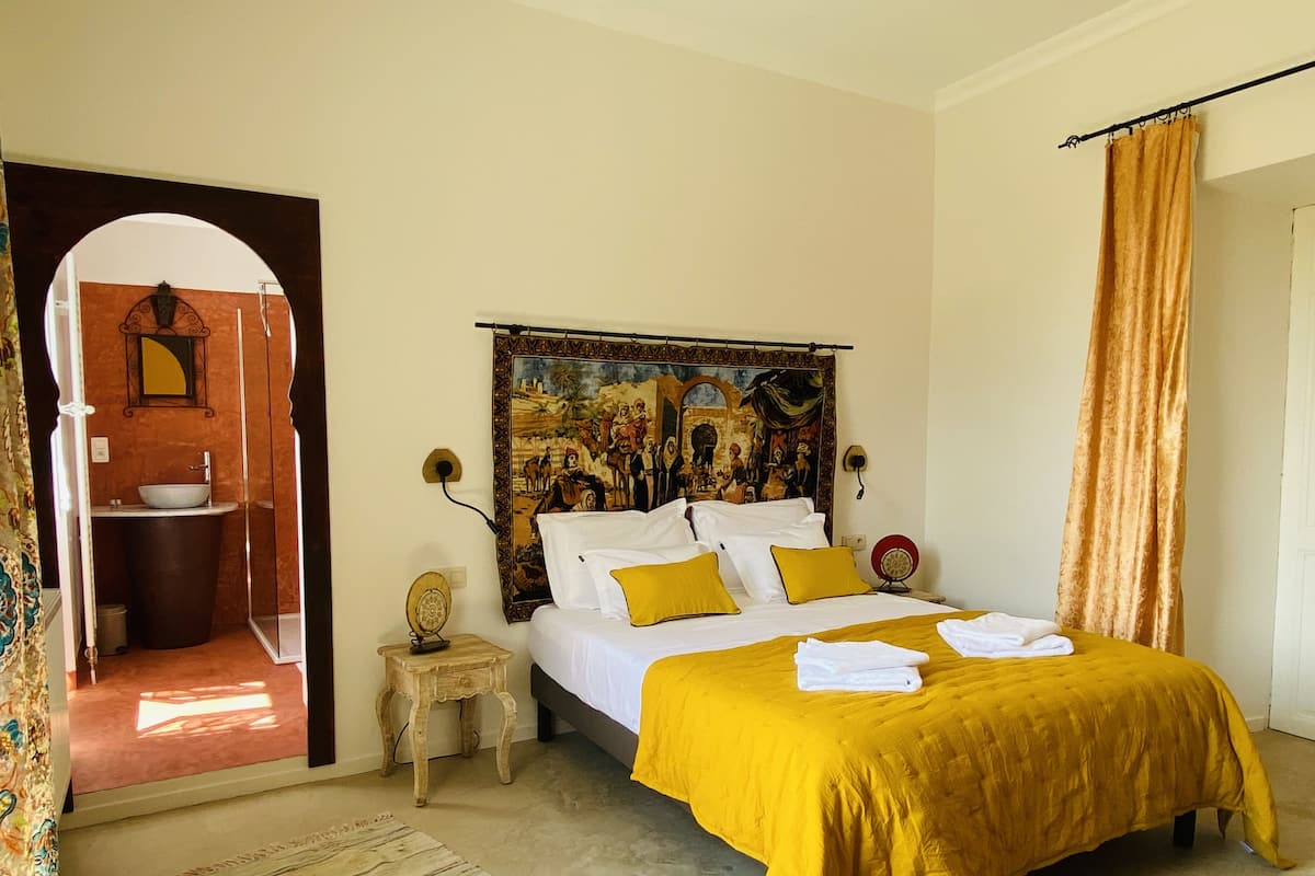A bedroom with a bed, a headboard with a mural, a small bedside table, a bathroom visible through an archway, and a curtain.
