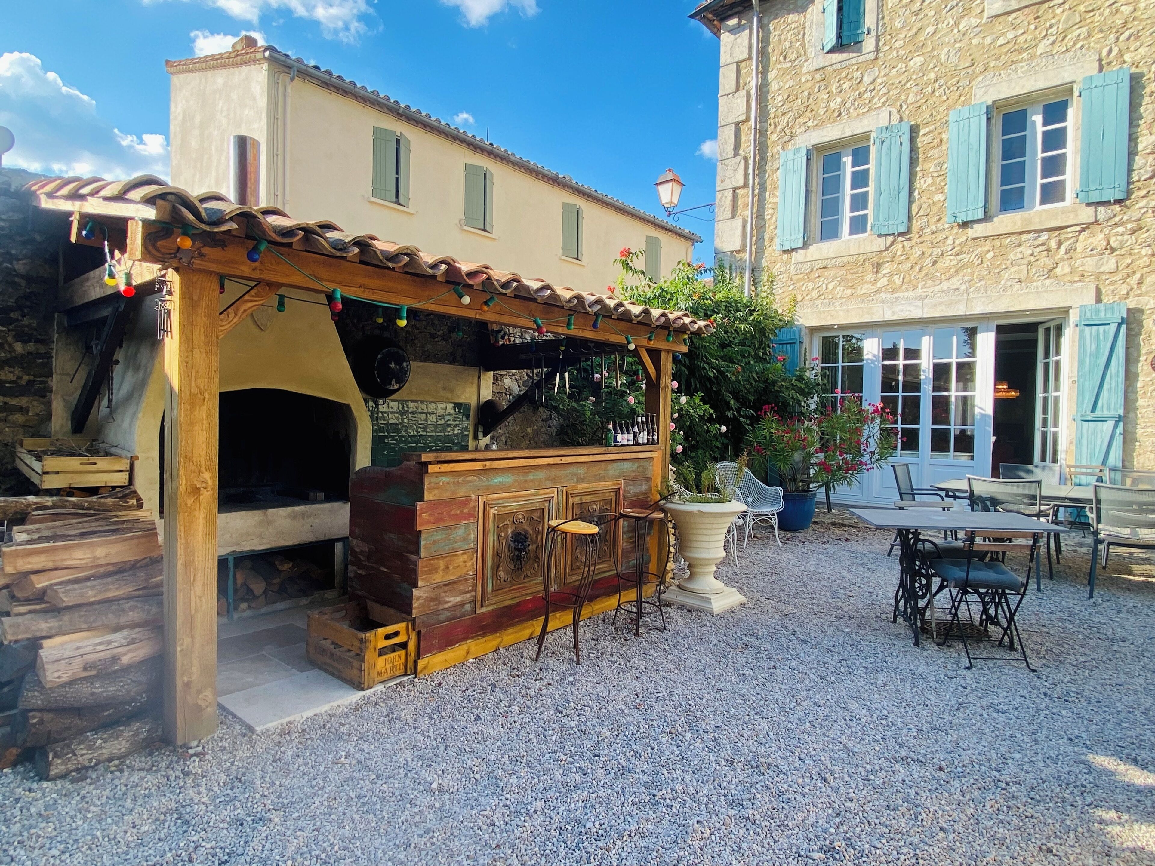 A rustic outdoor seating area with a wooden table and chairs, a stone building in the background, and a gravel ground.
