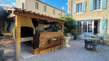 A rustic outdoor seating area with a wooden table and chairs, a stone building in the background, and a gravel ground.