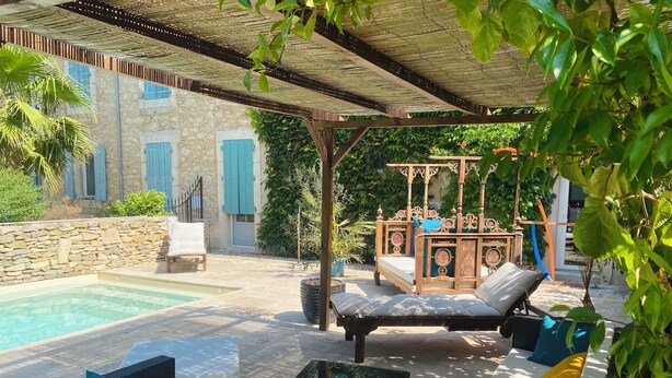 A poolside area with a covered seating area, a stone wall, and a view of a building with blue shutters. - Aux Berges du Libre (Félines-Termenès)