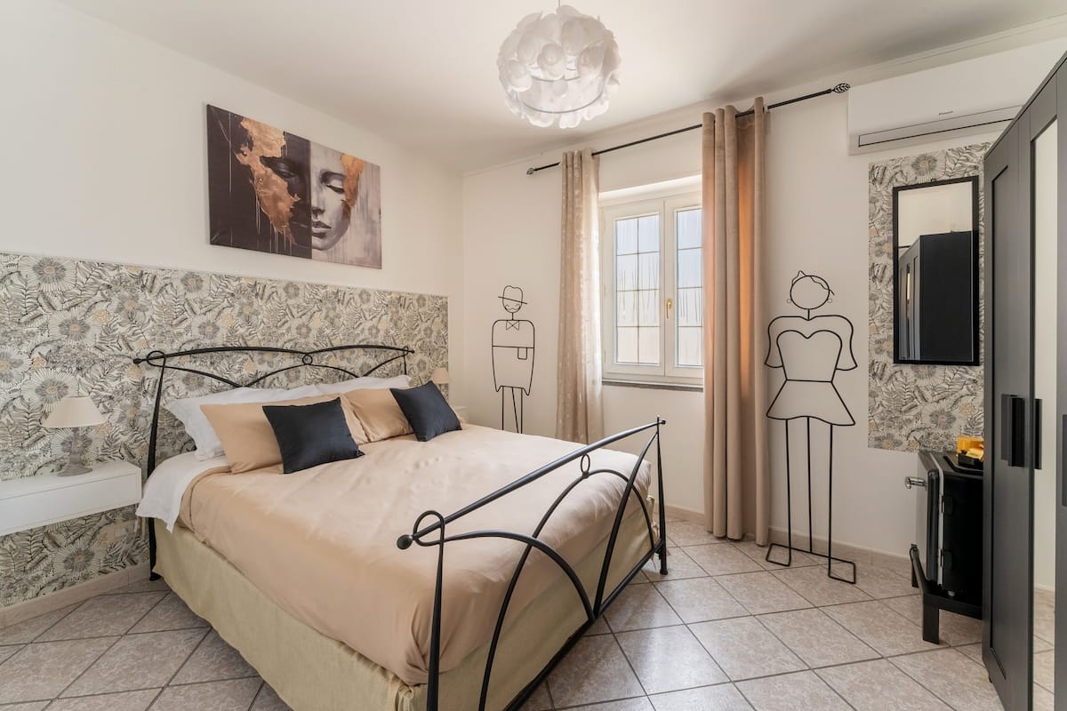 A bedroom with a bed, a nightstand, a wall-mounted lamp, a window with curtains, and a wall with a floral pattern.